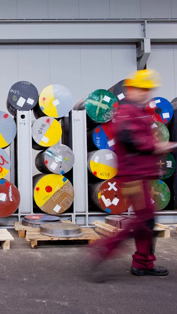 Foto: Gesco: Arbeiter in Schutzausruestung vor einer Wand mit bunten, gestapelten Metallspulen in einem Industriegelaende
